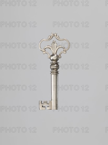 Furniture key, locksmithing; tubular handle with a rectangular quill with cutouts. Ball-shaped node separated from the decorative handle by two rings, and from the handle by a single ring with a wavy edge. Cast, openwork handle composed of channel sections with stylized leaves. Circa mid-18th century. Height 8.5 cm, width 3.5 cm, depth 1.1 cm. Maker, provenance unknown., A single metallic key is centered against a plain light gray background, featuring a decorative ornate bow with curved, symmetrical scrollwork and a rounded central knop, a fluted shaft with subtle ridges, and a rectangular bit with notches; the visible colors are silver metallic tones and shades of light gray.