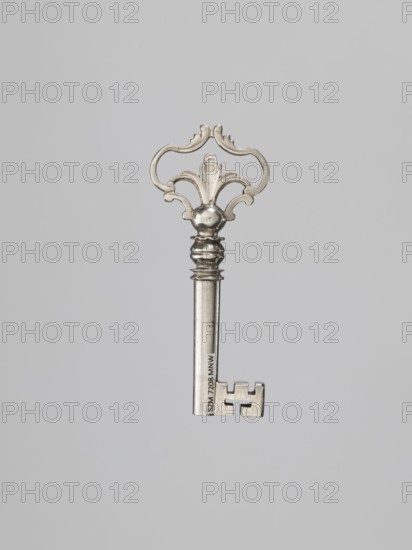 Furniture key, locksmithing; tubular handle with rectangular quill with cutouts. Ball-shaped node separated from decorative handle by two rings, and from the handle by a single wavy-edged ring. Cast openwork handle composed of channel sections with stylized leaves. Circa mid-18th century. Height 8.5 cm, width 3.5 cm, depth 1.1 cm. Maker and provenance unknown., A metallic ornamental key with a decorative bow showing a curved, symmetrical design and a central leaf-like motif, a rounded collar and a fluted shaft ending in a rectangular bit with stepped cutouts; the key is silver colored against a plain light gray background.