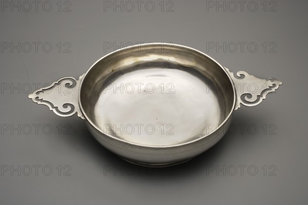 Écuelle by Christian Lieberkühn fl. 1703–1733, goldsmith. Hidden on the bottom: city mark not recorded in the literature, maker's mark Szeffler, pp. 58–59, no. 90. Circular, bulging, smooth vessel on a low base with two horizontal handles with openwork. Vessels of this type, appearing already in the 13th–14th centuries, were used for drinking broth and soups; they usually had a plate–stand and a cover topped with a handle or three legs, in the latter case serving as a stand. Unpublished. Berlin, Germany. Vessel; height 7 cm, width 25 cm, depth 15.5 cm, weight 333 g., A shallow round metal bowl with a smooth reflective interior and ornate flat handles extending from opposite sides, set on a flat gray surface; the bowl and handles appear silver in color with subtle darker and warmer metallic reflections, and the background is various shades of gray.