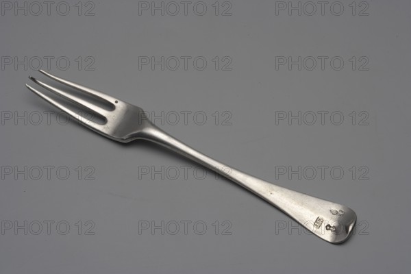 Three-pronged fork by Emmanuel I Ellerholtz active from 1758, died 1800, goldsmith, 1770. Three-pronged fork with a flat handle widening towards the end, ending in a semicircle and bent forward; on the reverse of the handle an engraved coat of arms under a crown. Materials: presumably silver/cutlery. Size: length 19 cm, width 2.2 cm, weight 55 g. Place: Gdansk, Royal Prussia Poland., A metal fork lies flat on a smooth gray surface. The fork has a long, tapered handle that widens slightly at the end and a three-pronged head with elongated spaces between the prongs. The metal shows a reflective, slightly worn finish with small scratches and subtle variations in tone. The background is uniformly gray with faint marks and smudges. Colors visible include silver from the fork and various shades of gray in the surface and background.