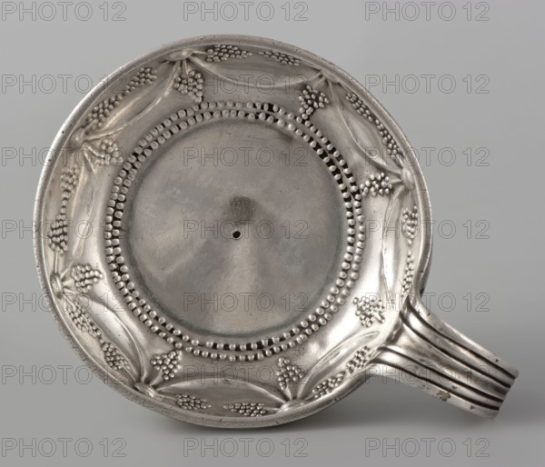 Bowl, Jean-Joseph X goldsmith, scrap - purchase, tableware element; bowl with engraved inscription on the side FRANCOIS PETIT, 1735 - 1737; Orleans, France; vessel; height 3.5 cm, width 10.8 cm, depth 8.5 cm., A round metal object with a shallow bowl and a short handle, shown from above, exhibiting a silvery-gray surface with polished and slightly tarnished areas; the rim features raised decorative motifs resembling small clustered beads and leaf-like shapes arranged around the circumference, and there is a dotted beaded ring encircling the central flat interior; the handle extends from one side with parallel ridged bands and shows similar silvery-gray tones with some darker spots.