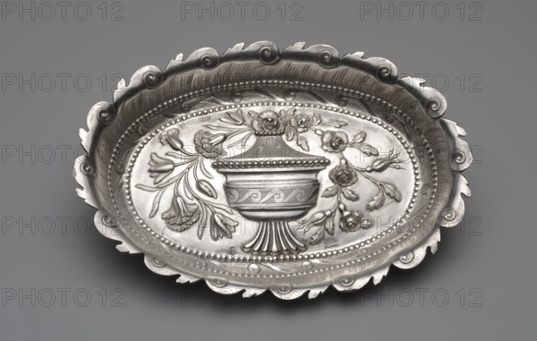 Platter by Johann Jacob Haase II active from 1759, died 1775, ca. 1780. Oval cake stand with a mirror decorated in relief with a covered vase topped by a bouquet of carnations, tulips, and roses; surrounded by a band of chien courant running wave motif enclosed in pearling, above it a rooster's comb; irregularly cut edge with a repeated running-wave motif. At the bottom a dotted inscription with the date MM & Fr.ACM / Ao 1782 4 Junii. Note: discrepancy between the goldsmith’s recorded activity died 1775 and the 1782 hallmark of Ellerholtz; studies do not confirm whether the widow or son continued the workshop. Silver; vessel. Measurements: height 2.2 cm, width 21.9 cm, depth 15.2 cm, weight 159 g. Made in Gdansk, Royal Prussia, Poland., A shallow decorative oval metal dish with an ornate scalloped rim and embossed floral and urn motifs, shown against a smooth gray background; the dish displays polished and matte areas with visible texture, a raised beaded inner border, stylized leaves ...