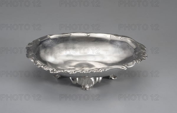 Bowl by Jacob Haase 1704–1762 or his widow; rocaille ornament, oval with a rocaille-decorated edge, supported by four leaf-shaped feet. Goldsmith's marks do not allow a more precise dating. The hallmark indicates it could have been hallmarked in 1752, 1758, or 1764; the goldsmith was deceased by 1764 but his widow ran the workshop until 1774. Dated to the 1750s or 1764. Gdansk, Royal Prussia, Poland. Vessel; height 2.6 cm, width 10.6 cm, depth 7.8 cm, weight 619 g., A shallow, oval metal bowl with a reflective silver surface sits on a smooth grey background; the bowl has an ornate raised rim with decorative scalloped edges and a few curved embellishments, and it rests on a small decorative base with rounded feet. The visible colors are silver and grey.