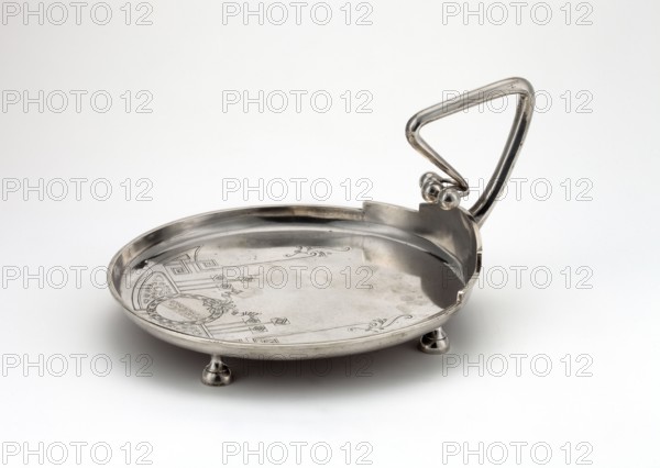 Silver circular plate with a handle on four legs, smooth edge; interior engraved with Art Nouveau geometric-floral ornament; broken handle imitating a branch topped with three spherical fruits on stems. Art Nouveau, geometric-floral ornament, 1908–1917, Moscow, Russia. Vessel. Height 13.7 cm; width 25.1 cm; depth 20 cm; weight 127 g. Maker, scrap—purchase, purchase, A shallow round metal dish with a reflective silver surface rests on a flat white background, supported by a few small rounded feet; the dish interior shows etched decorative patterns and a smooth area with subtle tarnish marks, and one side features a raised section leading to an angular, triangular metal handle attached with a pair of round knobs, all surfaces showing silver and gray tones with highlights from reflected light.