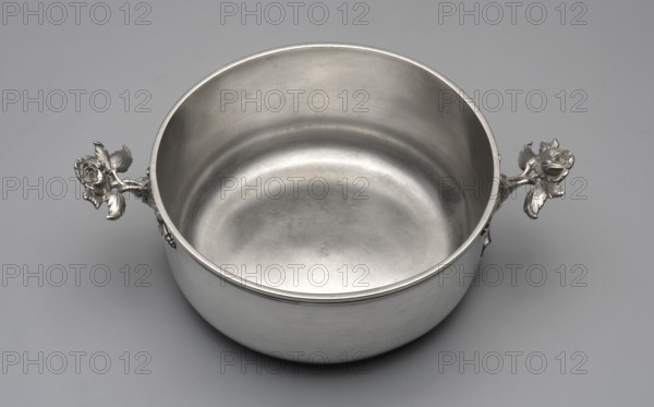 Vegetable pot, maker: Ephraim Wischcke fl. 1755–1799, goldsmith. Breischüssel type of vessel. Round vegetable pot with a flat bottom and profiled rim; handles formed as two plastic rose stems; no lid. Probably intended for serving boiled, chopped, or pureed vegetables hence called Breischüssel in German literature. Decoration: flower on stem rose. Date: 1770s. Place: Gdansk, Royal Prussia, Poland. Medium: vessel. Dimensions: height 7.4 cm, width 25 cm, depth 17.3 cm; weight 727 g., A metallic round bowl with a smooth interior and exterior surface sits on a flat background, showing a silver-gray sheen. The bowl has a simple rim and two decorative handles shaped like detailed flowers with leaves extending outward, also in silver-gray. The background is a uniform light gray. The overall visible colors are silver-gray and light gray.