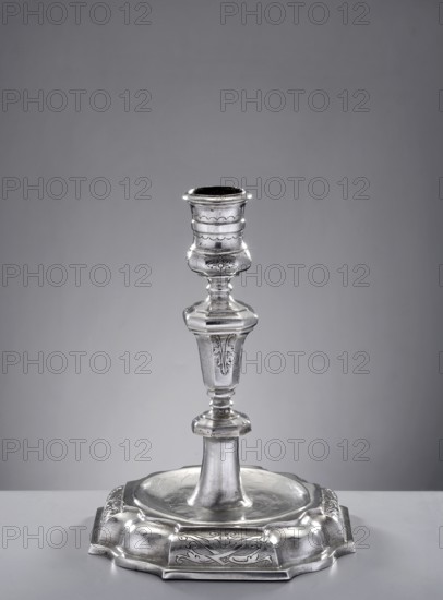 Single-candle candlestick by Hieronymus Holl II 1660–1732, goldsmith. On an octagonal foot with alternating straight sections, in the form of a half-cylinder with pincer and wavy decoration. The main surface is circular, slightly concave, from which the lower part of the shaft extends, terminating in an octagonal, flattened node. Above, the shaft is an octagonal baluster widening upwards; the sides are alternately smooth and engraved with acanthus leaves. Cylindrical profiled sleeve, expanded at upper and lower parts. Dated 1721–1730, made in Gdansk, Royal Prussia, Poland. Function: lighting. Height 14.5 cm; width 11.2 cm; weight 207 g., A single metallic candlestick stands centered on a plain surface against a neutral background, showing a shiny silver color with polished highlights and subtle darker tones in recessed areas; the candlestick has a flared base with decorative raised edges and engraved floral motifs, a tapered central stem with rounded and faceted sections, and a cup-lik...