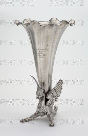 Vase by Grachev Brothers St. Petersburg; silversmith's factory; fl. 1866–1918. Vase in the form of a parted chalice with a profiled rim supported by a solid winged dragon; inscription on the chalice: On the day of the first confession / and holy communion / In memory of / Our beloved Elunia / Parents / 21 V 44. Before 1896, Russia, St. Petersburg. Vessel; height 34 cm, width 20 cm, weight 1787 g., A metallic vase with a flared, scalloped rim in a silvery tone sits on a sculpted base shaped like a winged creature; the creature has a textured body and detailed wings, a long neck, pointed ears or horns, and a fierce facial expression, all rendered in the same silvery color, with the vase surface showing a few subtle darker tarnish marks and reflections against a pale, light gray background.