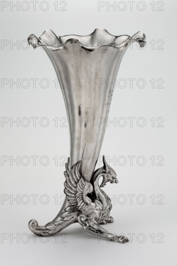 Vase, Grachev Brothers St. Petersburg; silversmith's factory; fl. 1866–1918; role: factory; inscription of factory/corporate slogans; scrap - purchase; dragons; dedications. Vase in the form of a parted chalice with a profiled rim supported by a solid winged dragon; inscription on the chalice: On the day of the first confession / and holy communion / In memory of / Our beloved Elunia / Parents / 21 V 44. Before 1896, Russia; St. Petersburg. Vessel; height 34 cm; width 20 cm; weight 1787 g., A silver-colored metal vase with a flared, ruffled rim sits on a light gray background, the tall body tapering down to an ornate base where a sculpted winged creature supports the vase; the creature has outstretched feathered wings, a long curved neck, a horned head, and muscular limbs that end in clawed feet, all rendered in the same metallic silver tone with subtle darker tarnish marks and reflective highlights.