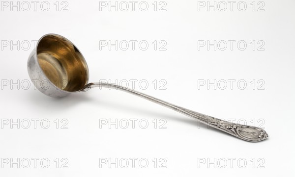 Vase spoon; Grachev Brothers silversmith factory St. Petersburg; active 1866–1918. Geometric-floral ornament; circular, flat-bottomed scoop, gilded inside. Widening handle ending in a semicircle with a point; handle decorated on both sides with relief interwoven ribbons and acanthus leaves; engraved RT ornament on the handle. Dated 1908–1917, Russia, Moscow. Cutlery; height 34 cm, width 9.4 cm., A metal ladle lies on a white surface, showing a rounded bowl with a golden-toned interior and a silver-toned exterior that has some darker spots; a long, slender silver-toned handle extends from the bowl and ends in an ornate, embossed decorative tip with raised patterns in silver tones.