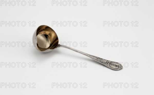 Sauce spoon by Grachev Brothers St. Petersburg; silversmith's factory; fl. 1866–1918. Geometric-floral ornament; hemispherical scoop with wide spout, gilded inside; widening handle ending in a semicircle with a point, decorated on both sides with relief interwoven ribbons and acanthus leaves; engraved RT ornament on the handle. Dated 1908–1917, Russia, Moscow. Cutlery. Height 17.8 cm, width 6.4 cm., A single metal ladle lies on a plain light background, showing a rounded bowl with a reflective interior that appears gold and a curved exterior that appears silver, attached to a long ornate handle that is silver with decorative raised patterns and a decorative oval end; the image includes shades of white, gold, silver, gray, and subtle darker reflections within the bowl.