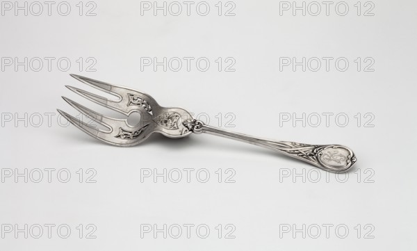 Fish platter fork by Grachev Brothers St. Petersburg; silversmith's factory; active 1866–1918. Four-pronged fish fork with floral decoration; widening handle ending in a semicircle with a tip, decorated on both sides with relief interwoven ribbons and acanthus leaves; engraved RT ornament on the handle. Geometric-floral ornament. Scrap-purchased, Dated 1908–1917, Moscow, Russia. Cutlery. Height 25.7 cm; width 5.6 cm., A metallic serving fork with an ornate handle rests on a light surface; the fork and handle are silver in color with darker engraved accents, featuring decorative cutouts and scrollwork on the head and a patterned, sculpted handle with floral-like motifs.