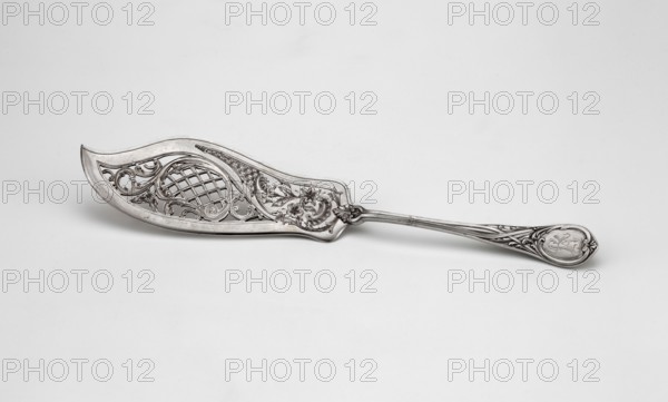 Fish platter, Grachev Brothers St. Petersburg; silversmith's factory; fl. 1866–1918 maker, scrap - purchase, geometric-floral ornament, openwork, with decoration of floral-rococo motifs; widening handle ending in a semicircle with a tip, decorated on both sides with a relief decoration of interwoven ribbons and acanthus leaves; engraved RT ornament on the handle, 1908–1917, Moscow, Russia; cutlery, table accessory; height 29.9 cm, width 5.9 cm, A single ornate metal utensil lies against a plain light background, showing a decorative broad blade with pierced latticework and scrolling floral motifs, connected to a long handle that has raised curving embellishments and a rounded end with an engraved cartouche; the visible colors are shades of silver, gray, and off-white.