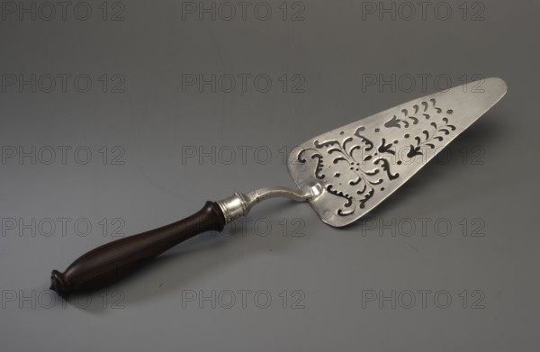 Cake server by Johann Christoph Conradi fl. ca. 1717–1742. Triangular with rounded corners, decorated with openwork in the form of plant tendrils and flower calyxes. The handle is curved, ending in a polygonal socket containing a wooden reconstructed handle. Typically this type of server is described in the literature as intended for fish, though the later typeification of shapes for fish, cakes, tarts, pâtés is noted. Dated 1717–1742, Berlin, Germany. Cutlery/table accessory. Height 22 cm; width 7.9 cm; weight 121 g., A metallic cake server with an ornate perforated triangular blade featuring decorative cutouts rests on a gray surface, attached to a curved metal neck and a dark brown wooden handle with a rounded end and subtle turned detailing; visible colors include silver, dark brown, and shades of gray.