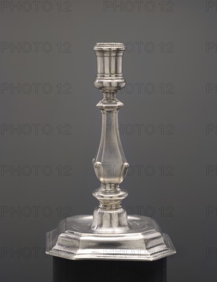 Candlestick, Spindler, Sebald fl. ca. 1716–1740, goldsmith. Candlestick on an octagonal profiled foot, baluster shaft narrowed towards the top, cylindrical candle socket. 1730s, Berlin, Germany. Candlestick lighting. Height 16.5 cm; width 10.3 cm; weight 292 g., A metallic candlestick with a polished silver finish stands against a smooth gray background; its design features a flared, ridged top leading to a bulbous neck, a tapered shaft with subtle curved panels and small projecting tabs, a rounded collar above a stepped, rounded base, and an octagonal lower platform with beveled edges resting on a dark support.