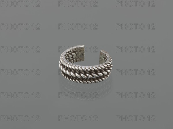 Bracelet; maker, 19th century, Middle East, Islamic art; body/female/hand ornament, fashion accessory; tugra motif; materials: jewelry unspecified; height 2.5 cm, width 7.0 cm, weight 170 g., A metallic ring with an open gap sits on a smooth surface, showing a braided rope-like texture around its exterior and a dotted, hammered pattern on its inner faces; the ring and its reflection display shades of silver and gray against a uniform gray background.