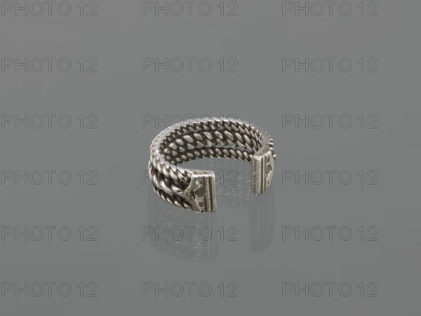 Bracelet; maker, 19th century; Middle East possibly; Islamic art; body ornament—female hand ornament; tugra motif; jewelry; materials unspecified; height 2.5 cm; width 7.0 cm; weight 170 g., A small metal ring with a braided design forming several intertwined rows, open at one side with two flat end caps that have textured surfaces; the ring and its reflection rest on a smooth surface. Visible colors include silver, gray, and tonal variations of those shades.