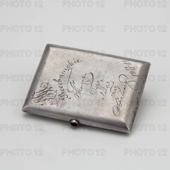 Cigarette case, scrap metal, Art Nouveau style, decorated with bats. Rectangular cigarette case with rounded corners and a decoration on the lid in the shape of a flying bat. On the reverse, an engraved tied Cyrillic monogram WAP and a facsimile of donors' signatures and the date 29 June 1913. Early 20th century, Russia. Smoking accessory. Height 7.5 cm, width 10.1 cm, weight 160 g., A rectangular metallic case with rounded corners lies on a pale surface; the case has a slightly raised rim and a small rounded clasp on one long edge. The metal shows a worn, scratched texture with areas of darker tarnish and lighter highlights, appearing primarily silver and gray. On the top surface there are decorative engraved flourishes and flowing script-like incisions forming curved lines and loops; the engraving contrasts as darker grooves against the metal. The overall scene includes a few subtle shadows beneath the case and a clean, neutral background in shades of off-white and very light gray.