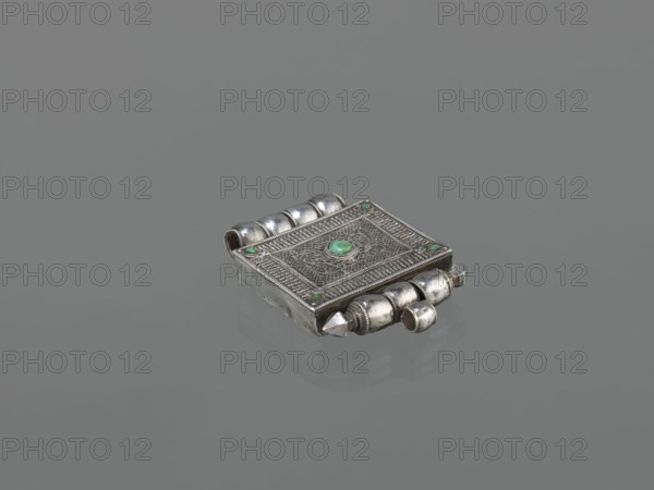 Ga'u box fragment, maker, plant twig; jewelry; Himalayan culture; Tibetan Buddhist art; amulets; Tibetan culture; provenance, 19th century; Tibet; varia; height 11.0 cm; width 7.5 cm; weight 125 g, A small ornate rectangular metal box sits on a smooth gray surface, its body displaying detailed raised filigree and patterned borders. The metal has a tarnished silver appearance with areas of darker patina, and a few small rounded green stones are set into the top and corners. One side of the box features a series of rounded tubular shapes forming a decorative hinge-like row, and a rounded knob projects from one end. The top surface shows concentric decorative panels and embossed motifs, while the underside and sides retain the same silvery metal with subtle reflections. The background and surface are uniform gray.