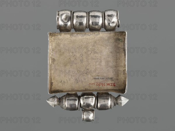 Ga'u box fragment, maker, plant twig, jewelry; Himalayan/Tibetan Buddhist amulet; Tibetan culture; 19th century, Tibet; varia; height 11.0 cm, width 7.5 cm, weight 125 g, A rectangular metal object with a worn, beige-toned interior surface set against a gray background, flanked on the top and bottom by a group of rounded, silver-colored bead-like metal elements with beaded edges and a slightly hammered texture, and with pointed, faceted silver-colored caps at the ends of the lower group.