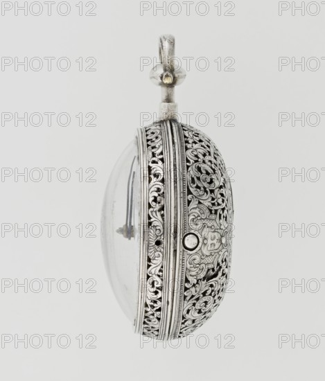 Pocket watch by Carl Willougby, 2nd quarter of the 18th century, London, England. Silver pocket watch with striking function in a partially openwork case, richly engraved over the surface with acanthus twigs, flowers, birds, two masks male and female, acanthus scrolls, fish-scale ornament, shells and decorative masks. Center features a circular reserve of acanthus twig, volutes and fish scales enclosing a bell. Glazed lid set in an openwork frame decorated with a floral twig and a broken ribbon. Mechanism between two circular plates supported by columns, with spindle escapement, a worm with chain, and a balance drum. Gilded openwork crown engraved with an acanthus twig, combined with an openwork foot and a woman's mask. White-enameled dial edged with a gilded knurled ribbon, Arabic hour numerals, minutes marked with vertical lines; steel hands. Height 8 cm; width 5.5 cm; dial diameter 4.5 cm., A decorative metallic oval object with ornate filigree patterns in silver tones on one side a...