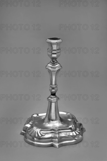 Candlestick by Johann Gottfried Schlaubitz fl. 1733–1771, goldsmith. Scrap - purchase, purchase provenance. On a quatrefoil foot, arched and profiled. In the center of the segments are alternating shells and volutes with a fan motif. From the slightly concave surface a conical lower part of the shaft protrudes, upon which a baluster with octagonal profiles is placed. Vase sleeve with octagonal profiles. Dated 1740s. Origin: Royal Prussia, Gdansk, Poland. Function: candlestick lighting. Dimensions: height 19.4 cm; width 13.1 cm; depth 13.2 cm. Weight: 295 g., A single metallic candlestick stands upright against a plain background, showing a polished reflective surface in shades of gray and silver. The base is ornate with curved, scalloped edges and flowing, embossed designs that catch highlights and shadows. The stem tapers upward with a series of decorative, faceted sections and rounded nodes, each reflecting light differently to create contrasts of brighter silver and darker gray. The...