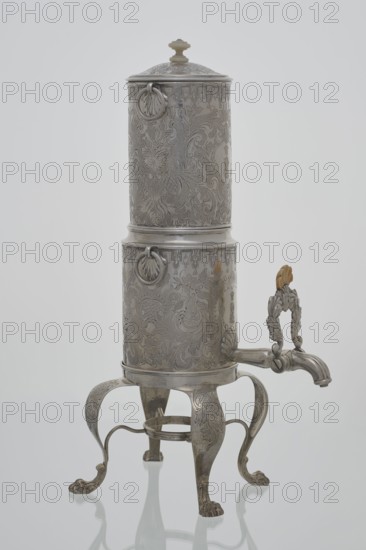 Coffee maker by Stephan Mayerhofer fl. 1818–1852, goldsmith; 1835, Vienna, Austria. Vessel with lion’s paw feet; height 35 cm, width 15 cm, depth 19 cm; weight 960 g., A tall ornate metal vessel with a cylindrical body and a domed lid sits on a decorative four-legged stand; the vessel and stand have intricate engraved floral and scrollwork patterns all over their surfaces, and there are a few circular ring handles attached near the top and middle of the cylinder. The stand's legs curve outward and end in clawed feet, and a curved spout extends from the lower portion of the cylinder with a decorative handle above it that features leaf-like motifs and a small carved element in a light tan color. The overall colors visible are silvery metal tones, light tan on the small carved element, and subtle reflections of white and soft gray from the background.