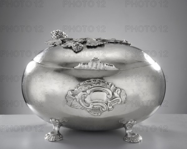 Rococo sugar bowl by Emmanuel I Ellerholtz fl. 1758–1800, goldsmith. Ovoid sugar bowl supported on four bent shell legs; front has a relief rocaille blind reserve. Hinged lid with a handle in the form of a rooster’s comb, topped with a modeled vine branch with clusters. Materials/ornaments: rocaille, vine, rooster’s comb. Style: Rococo. Date: 1768, Gdansk, Royal Prussia, Poland. Function: sugar vessel. Dimensions: height 10 cm, width 14.5 cm, depth 10 cm; weight 385 g. Provenance: purchase., A shiny metallic oval container with an ornate lid sits on a flat surface supported by a few short decorative feet; the surface of the container is reflective and smooth with embossed ornamental detailing on its front and floral-like embellishments on the lid. The colors visible are shades of silver and gray across the container and lid, with lighter silver highlights where light reflects and darker gray shadows on the underside and background. The background is a gradient of gray.