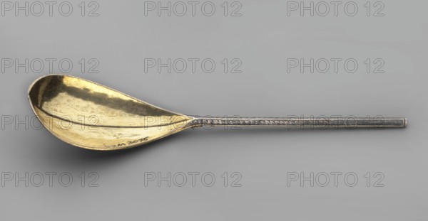 Baroque spoon, baroque style, plant ornament, pearling, cutlery. Baroque spoon with a teardrop-shaped scoop, broken along the axis, gilded inside; top decorated with a niello ornament of plant tendrils in radial reserves; angular handle decorated with scales, pearling and small leaves. Late 17th century, Poland. Height 23 cm, width 5 cm., A metal spoon with a long handle and a shallow, oval bowl lies horizontally against a light gray background; the bowl has a warm golden color with darker tarnished areas and subtle scratches, while the handle has a cooler silvery-gray tone with a decorative raised edge and slight patina.