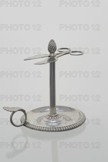 Scissor-shaped candlestick stoczek, Jan Radecki fl. 1815–1837, goldsmith. Candlestick for a thin candle twisted serpentine around the stem; supported on a plate-shaped base edged with a half-cylinder; provided with a handle in the form of a ring with a thumb rest. Stem is a sleeve around which the thin candle is twisted; sleeve topped with a pine-cone finial with a vice in the form of flat scissors with volute handles to extend the candle to the desired length. 1815–1837, Poznan, Poland. Function: lighting. Dimensions: height 14.5 cm, width 14 cm, depth 11 cm., A metallic object with a circular base and a raised central column supporting a horizontal pair of scissor-like blades and two circular loops; the base has a ridged decorative rim and a shallow dish center, with a ring-shaped handle attached to the rim; the top of the column is topped with an ornamental, textured finial. Visible colors include silver, gray, and reflections of pale white.