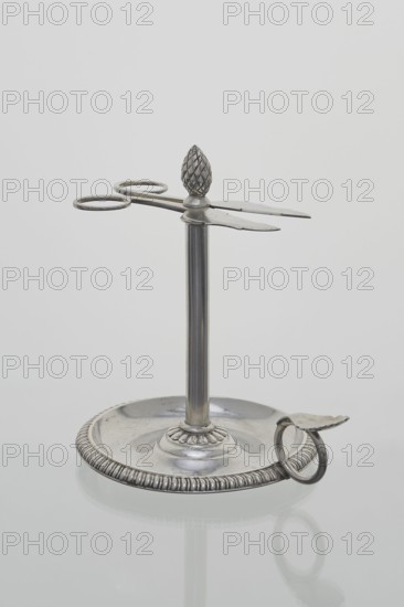 Scissor-shaped candlestick stoczek, Jan Radecki fl. 1815–1837, goldsmith. Candlestick for a thin candle that was serpentine-twisted around the stem; supported on a plate-shaped base edged with a half-cylinder and provided with a ring handle with thumb rest. Stem formed as a sleeve around which the thin candle twisted serpentine; sleeve topped with a pine-cone finial with a vice in the form of flat scissors with volute handles to extend the candle to desired length. 1815–1837, Poznan, Poland. Function: lighting. Height 14.5 cm; width 14 cm; depth 11 cm., A metallic stand with a circular base and a vertical central rod supporting a horizontal scissor-like element topped by a textured finial shaped like a pinecone; the base has a ridged outer rim and a small curved handle attached at the edge, and the object is shown against a plain light gray background. The visible colors are silver and shades of gray.