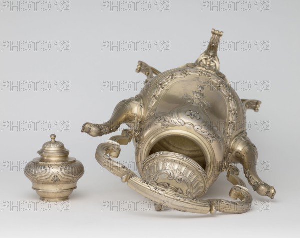 Kettle with warmer; maker: André Aucoc, goldsmith; tableware; kettles; 1887–1911; France, Paris; vessel; height 39 cm, width 27 cm, depth 17 cm, A metallic ornate vessel lying on its side next to a separate lid, both showing elaborate raised floral and scrollwork decoration in a metallic gold-silver tone; the vessel has curved handles and claw-like feet with decorative detailing, and the interior visible through the opening shows a similar metallic finish; the background is a smooth light gray.