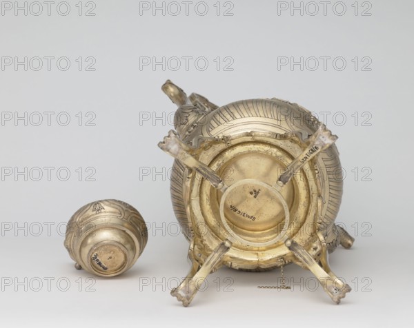 Kettle with warmer, Aucoc, André goldsmith, tableware items, kettles, 1887–1911, France, Paris; vessel; height 39 cm, width 27 cm, depth 17 cm, A large ornate metal vessel lies on its side showing its rounded base and four decorative curved legs extending outward, with a small chain hanging from the rim; nearby is a smaller rounded metal lid or cup placed on its side. The metal surfaces display a polished golden tone with areas of darker tarnish and painterly patina, and there are engraved decorative patterns and fluted ridges on the larger piece and similar ornamental motifs on the smaller piece. The background is a plain, light neutral color.