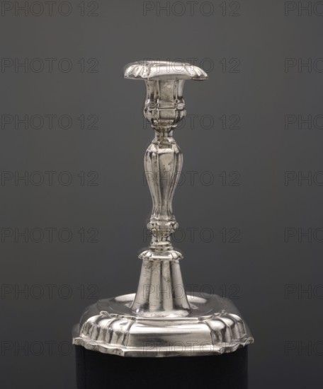 Candlestick, Junge, Andreas I fl. 1710–1757 goldsmith; single-candle candlestick, smooth on a multipartite, slightly arched, profiled base; baluster shaft, angular socket with profiled, — dated 1755; Royal Prussia, Königsberg; function: lighting; height 15 cm, width 11 cm, A metallic candlestick with a reflective silver surface stands against a uniform dark gray background; it has an ornate, curving central column with rounded and fluted sections, a decorative top platform, and a broad decorative base with scalloped edges, all showing highlights and slight tarnish.