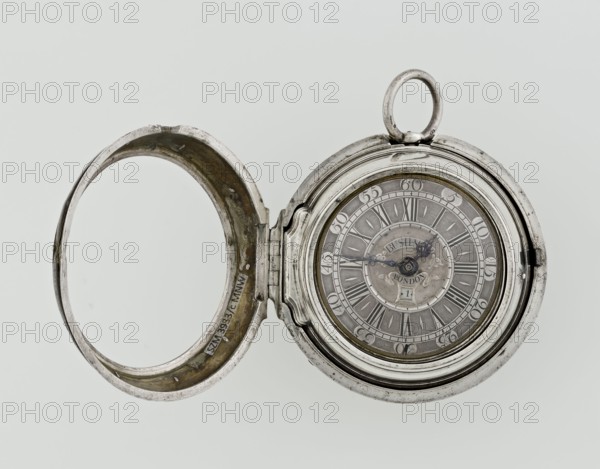Pocket watch by John Bushman 1692–1720. Silver pocket watch in three cases: outer case covered with leather, rim decorated with studded silver rosettes; middle and inner cases plain. Underside of inner case stamped with casemaker's initial H and number 3081. Transparent lid. Silver champlevé dial with Roman hour markers and Arabic minute markers in oval reserves on a matte background; central circular reserve with the watchmaker's signature on two semicircular plates reading BUSHMAN and LONDON; date window at the bottom. Steel hands with openwork hour hand. Movement with plate skeleton, spring drive, worm gear, and Gall chain; spindle escapement. Gilded lower plate with an openwork English-style bun in the form of a wreath filled with acanthus tendrils and an openwork grid, connected by a mascaron mask with an openwork bun foot. Hair adjustment is a rotating silver-plated dial with a scale I–VI. Engraved on the plate in italics: Jn Bushman 3081 / LONDON. Circa 1700, London, England. Ti...