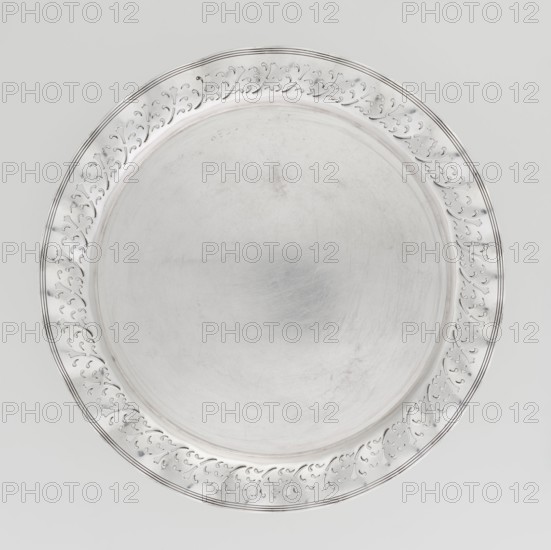 Circular tray, scrap - purchased, ca. 1908, England, Birmingham; dish; diameter 32 cm; weight 722 g, A round metallic tray with a reflective silver surface fills the image, showing a smooth central area with light scratches and subtle circular marks, surrounded by a raised decorative rim that features a repeating carved vine-like pattern and thin concentric grooves; the overall colors visible are silver and shades of gray against a light gray background.