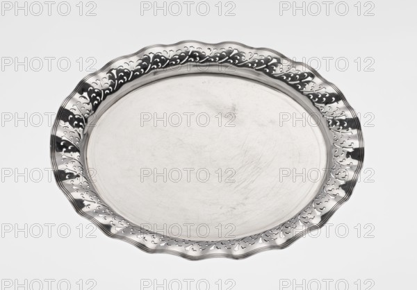 Circular tray, scrap, purchased, ca. 1908, England, Birmingham; dish; diameter 32 cm; weight 722 g, A round metal tray with a smooth central surface showing faint scratches and scuffs, surrounded by a decorative scalloped rim with cutout ornamental patterns; the tray and rim appear in reflective silver and gray tones set against a plain light gray background.