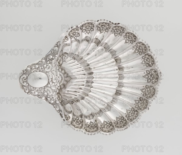 Shell-shaped silver platter by Atkin Brothers Silversmith Ltd Sheffield; goldsmith and silversmith firm; 1826–1958, scrap–purchase provenance; floral ornaments, scallop-shell shape, acanthus leaves ornament, clasp, relief decorations. Silver platter in the shape of a scallop shell, terminating on one side with an oval narrow handle, radial clasp, decorated on the rim with relief floral ornament, volutes and acanthus scrolls, supported on three shell feet. Dated 1893/1894, Sheffield, England. Vessel; height 5 cm, width 10 cm handle?, overall 28.5 × 23 cm; weight 357 g., A decorative shell-shaped dish in metallic silver tones fills the frame, showing a scalloped rim and radiating ridges that create a fan-like pattern; the surface features ornate raised floral and scrollwork embellishments in darker and lighter shades of silver, with areas of subtle shine and shadow that emphasize the embossed textures, and an oval smooth cartouche near the narrower end surrounded by intricate curling mot...