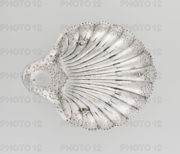 Shell-shaped platter by Atkin Brothers Silversmith Ltd Sheffield; goldsmith and silversmith firm; 1826–1958, silver, 1893–1894. Scallop-shell form terminating on one side with an oval narrow handle; radially clasped; rim decorated with relief floral ornaments, volutes and acanthus scrolls; supported on three shell feet. Floral ornaments; acanthus leaves. Vessel. Height 5 cm; depth 23 cm; width 28.5 cm; handle width 10 cm; weight 357 g., A silver, shell-shaped object with a scalloped edge and radiating ridges, showing decorative punched and engraved patterns and some dark tarnished spots, set against a pale gray background.
