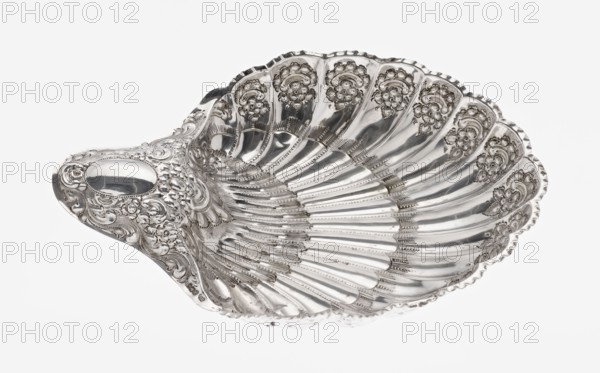 Shell-shaped silver platter by Atkin Brothers Silversmith Ltd Sheffield; goldsmith and silversmith firm, 1826–1958, scrap—purchase; floral ornaments; scallop-shaped shell with acanthus leaf ornament, clasp, relief decorations. Silver platter in the shape of a scallop shell terminating on one side with an oval narrow handle, radially clasped, decorated on the rim with relief floral ornament, volutes and acanthus scrolls; supported on three shell feet. Dated 1893–1894, Sheffield, England. Vessel. Height 5 cm; handle width 10 cm; overall width 28.5 cm; depth 23 cm; weight 357 g., A decorative shell-shaped dish with ornate raised floral and scrollwork patterns, showing a glossy reflective surface in silver tones; the dish has radiating ridges and scalloped edges with detailed embossed motifs, and a rounded handle-like extension with intricate embellishments, all photographed against a plain white background with visible highlights and reflections.