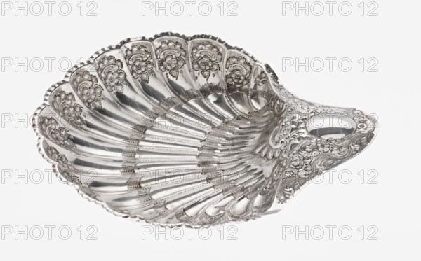 Shell-shaped silver platter by Atkin Brothers Silversmith Ltd Sheffield; goldsmith and silversmith firm; 1826–1958, 1893–1894. Scallop-shaped vessel terminating on one side with an oval, narrow handle; radially clasped; decorated on the rim with relief floral ornament, volutes and acanthus scrolls; supported on three shell feet. Materials: silver. Subject/ornament: floral ornaments, acanthus leaves, scallop shell motif. Dimensions: height 5 cm, depth 23 cm, width 28.5 cm, handle width 10 cm; weight 357 g., A decorative shell-shaped metal dish with a shiny silver surface, ornate raised floral and scrollwork patterns along the ridged interior and outer rim, a scalloped edge, and a smooth oval area near one end; colors visible include silver and shades of gray against a white background.