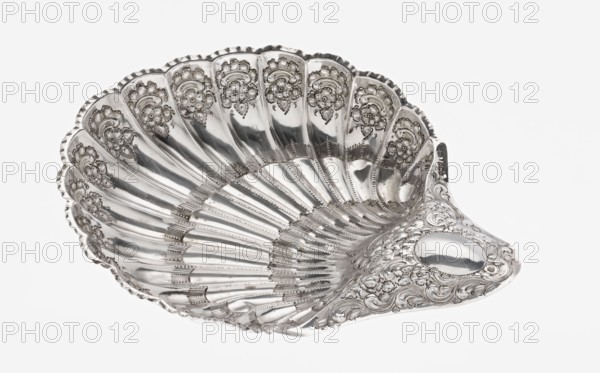Shell-shaped silver platter by Atkin Brothers Silversmith Ltd Sheffield; goldsmith and silversmith firm; 1826–1958, 1893–1894. Silver platter in the shape of a scallop shell, terminating on one side with an oval narrow handle, radial clasp; rim decorated with relief floral ornament, volutes and acanthus scrolls; supported on three shell feet. Floral ornaments; acanthus leaves; scallop shape. Vessel. Height 5 cm; width handle? 10 cm; length 28.5 cm; depth 23 cm; weight 357 g. England, Sheffield., A decorative silver shell-shaped dish with a shiny metallic surface and ornate embossed floral and scrollwork patterns along the scalloped rim and the inner curved sections, showing a series of radiating ridges and grooves that follow the shell form; the metal reflects light creating areas of bright highlights and darker gray reflections, and the overall colors visible are silver and shades of gray against a white background.