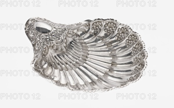 Shell-shaped silver platter by Atkin Brothers Silversmith Ltd Sheffield; goldsmith and silversmith firm; 1826–1958, 1893–1894. Scallop-shaped vessel terminating on one side with an oval narrow handle and radial clasp; decorated on the rim with relief floral ornament, volutes and acanthus scrolls; supported on three shell feet. Floral ornaments; acanthus leaves; relief decorations. Height 5 cm, depth 23 cm, width 28.5 cm, alternate width 10 cm, weight 357 g., A decorative shell-shaped metal dish with ornate raised floral and scrollwork detailing and a highly reflective silver surface, showing a few smooth, curved ridges radiating from the center and several scalloped edges, photographed against a plain white background with visible highlights and reflections.