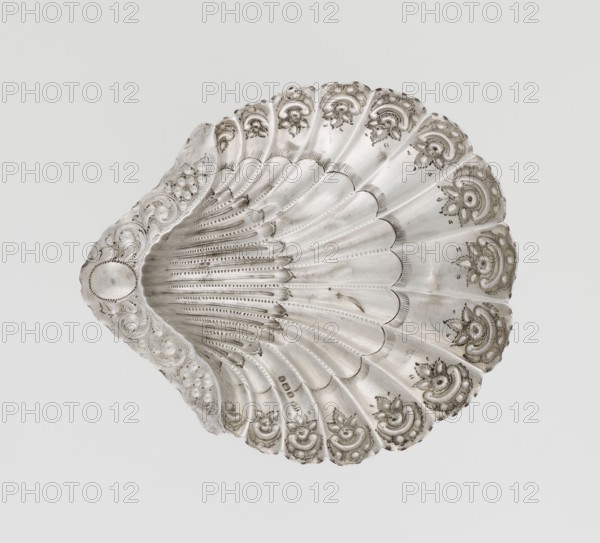 Platter in the form of a shell, Atkin Brothers Silversmith Ltd Sheffield; goldsmith and silversmith firm; 1826–1958, silver platter in the form of a full-plastic scallop shell, knotted and ribbed, rim decorated with a motif of relief flowers, leaves and channel sections; supported on three shell feet, 1890/1891, Sheffield, England, Great Britain; vessel; height 2.5 cm, width 15.3 cm, depth 13.1 cm, weight 113 g, A decorative shell-shaped object with intricate carved and embossed patterns, showing scalloped ridges radiating from a curved center and a border of repeating ornamental motifs; the piece appears metallic with a reflective silver color and areas of darker gray patina, set against a plain light gray background.