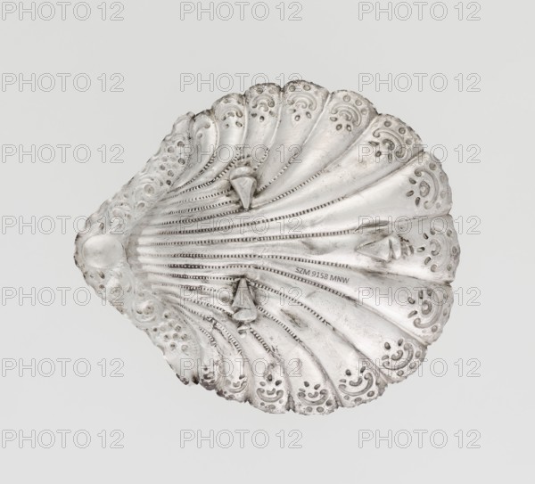 Platter in the form of a full-plastic scallop shell, knotted and ribbed, decorated on the rim with a motif of relief flowers and leaves and channel sections; supported on three shell feet. Silver. Maker: Atkin Brothers Silversmith Ltd Sheffield, c. 1890–1891. Dimensions: height 2.5 cm; width 15.3 cm; depth 13.1 cm; weight 113 g., A decorative scallop-shaped object with a metallic silver surface, showing a symmetrical fan of ridged segments radiating from a rounded end; each segment has ornate punched and embossed patterns with scalloped edges and tear-drop motifs, and there are a few raised cone-like studs along the ridges. The colors visible are primarily silver with areas of darker gray patina and subtle shadows against a light gray background.
