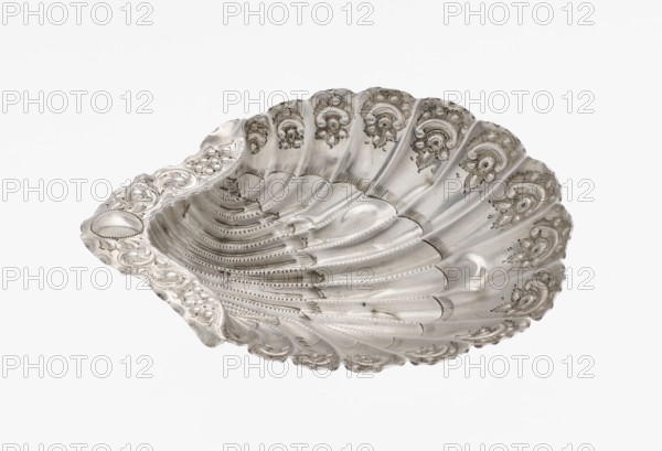 Platter in the form of a shell, Atkin Brothers Silversmith Ltd Sheffield; goldsmith and silversmith firm; 1826–1958 maker. Silver platter in the form of a full-plastic scallop shell, knotted and ribbed, decorated on the rim with a motif of relief flowers, leaves and channel sections; supported on three shell feet. Floral ornaments; shell shape; knotting; scallop shell. 1890/1891, Sheffield, England, Great Britain. Vessel; height 2.5 cm, width 15.3 cm, depth 13.1 cm, weight 113 g., A silver-colored, shell-shaped dish with a scalloped edge and ornate raised floral and scrollwork decorations around the rim, showing a textured, ribbed interior with repetitive curved grooves and dotted patterns, viewed against a plain white background.
