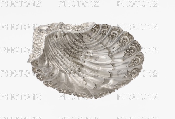 Platter in the form of a shell, Atkin Brothers Silversmith Ltd Sheffield; goldsmith and silversmith firm; 1826–1958 maker, scrap - purchase, floral ornaments, floral ornament, shell shape, knotting, scallop shell. Silver platter in the form of a full-plastic scallop shell, knotted and ribbed, decorated on the rim with a motif of relief flowers, leaves and channel sections; supported on three shell feet, 1890/1891, Sheffield, England, Great Britain. Vessel; height 2.5 cm, width 15.3 cm, depth 13.1 cm, weight 113 g., A scallop-shaped metallic dish with an ornate rim and a textured, ribbed interior, displaying decorative scrollwork and floral-like motifs along the edges; the surface is reflective with polished and matte areas, showing highlights and subtle shadowing; visible colors include silver tones, grey, and hints of darker tarnished grey.