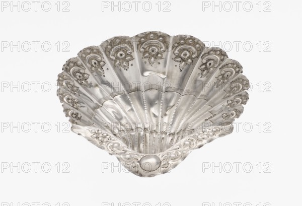 Platter in the form of a shell, Atkin Brothers Silversmith Ltd Sheffield; goldsmith and silversmith firm; 1826–1958 — Silver platter in the form of a full-plastic scallop shell, knotted and ribbed, decorated on the rim with a motif of relief flowers, leaves and channel sections; supported on three shell feet; floral ornaments; shell shape; knotting; scallop shell; vessel; 1890/1891; Sheffield, England, Great Britain; height 2.5 cm, width 15.3 cm, depth 13.1 cm, weight 113 g., A decorative shell-shaped object with a metallic silver surface, showing a scalloped fan form with fluted ridges and embossed floral and scroll motifs along the rim and inner panels, a smooth reflective finish with areas of subtle patina, and a background of plain white.