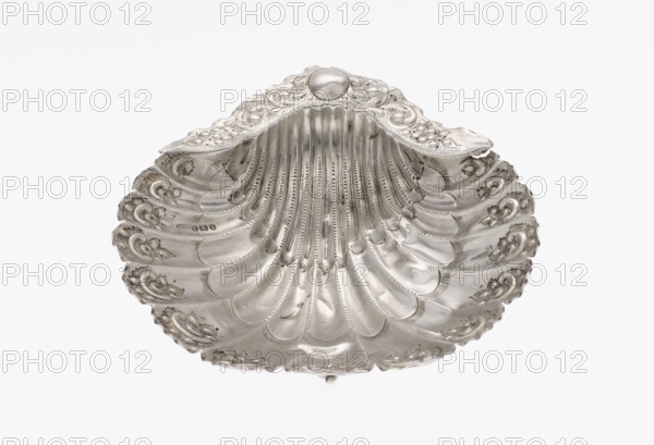 Platter in the form of a full-plastic scallop shell, knotted and ribbed, decorated on the rim with a motif of relief flowers and leaves and channel sections; supported on three shell feet. Maker: Atkin Brothers Silversmith Ltd Sheffield; goldsmith and silversmith firm; active 1826–1958. Date: 1890/1891. Place: Sheffield, England. Materials: silver. Function: vessel/platter. Dimensions: height 2.5 cm; width 15.3 cm; depth 13.1 cm; weight 113 g. Features: floral ornaments; shell shape; scallop shell; knotting; ribbing. Provenance: scrap — purchase., A single ornate shell-shaped object fills the frame, showing curved ridges and scalloped edges with embossed decorative motifs. The surface appears metallic with a polished sheen, reflecting light in gradients from bright highlights to softer gray tones. The object features repeated fan-like grooves radiating from the top center and small raised ornamental swirls along the rim. The overall colors visible are silver and varying shades of gray ...