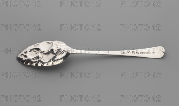 Jam spoon by Stephen I Adams fl. ca. 1758–1802. Jam spoon with an ovoid scoop with an irregularly cut edge, decorated with repoussé fruit on a leafy branch; smooth, widening handle terminating in a semicircle and bent inward at the end. Repoussé decoration from the mid-19th century ca. 1800/1801 noted; repousse decoration mid-19th century. London, England, Great Britain. Cutlery; length 22 cm, width 4.5 cm dipper, weight 62 g., A single metallic spoon lies horizontally on a gray background, showing a reflective silver surface with darker shadowed areas; the bowl of the spoon has a textured, embossed pattern with irregular raised shapes and darker recesses, while the handle is smooth and tapers slightly toward a rounded end with a subtle decorative curve; overall visible colors are silver, gray, and blackish shadows.