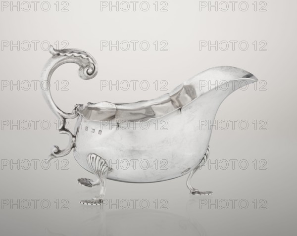Sauce boat; maker: Henry Matthews fl. 1893–ca. 1930, goldsmith; hemispherical silver sauce boat with an elongated wide spout and irregularly cut rim; supported by three palmette feet; volute handle profiled and decorated with an acanthus leaf; ornament: acanthus, volutes, palmettes; dated 1903–1904; place: Birmingham, England, Great Britain; vessel; height 11.5 cm, width 16 cm, depth 9.7 cm, weight 248 g., A silver-colored metal gravy boat with a high curved spout and an ornate scrolled handle, shown against a pale neutral background that transitions from off-white to light gray. The body of the vessel has a polished but slightly worn surface with visible reflections and subtle surface marks. The rim is irregular and slightly ruffled, and the interior shows a reflective silver finish. It stands on a few decorative feet resembling stylized shells or scrolls, and there is a hint of darker tarnish in recessed areas and along some edges. The colors visible are silver, off-white, and light ...